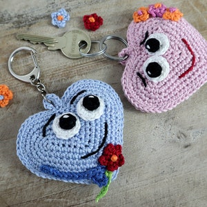 以下が含まれることがあります： 笑顔と大きな目で、2つのハート型の編みぐるみキーホルダー。1つはオレンジ色の花が付いたピンク、もう1つは赤い花が付いたブルーです。