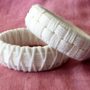 Könnte beinhalten: Zwei weiße geflochtene Armreifen, einer etwas größer als der andere, auf einem rosa Stoffhintergrund.