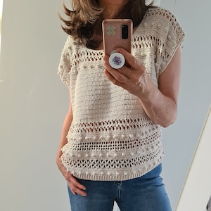 Peut inclure: Un haut en crochet beige avec un motif ajouré délicat et une rangée de petites pompons blancs autour du bord inférieur. Le haut a des manches courtes et un décolleté arrondi.