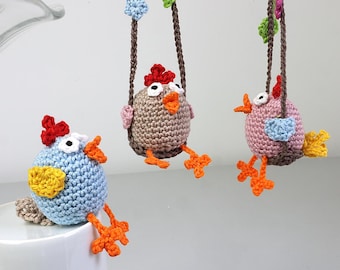 May include: Three crocheted chicks hanging from swings. The chicks are brown, pink, and blue with yellow beaks and orange feet.