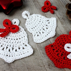 Könnte beinhalten: Drei gehäkelte Weihnachtsglocken, zwei weiße mit roten Schleifen und eine rote mit einer weißen Schleife. Die Glocken liegen auf einer Holzoberfläche.