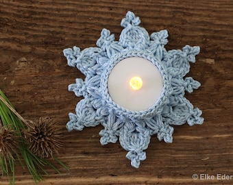 May include: A light blue crocheted snowflake candle holder with a white tea light candle inside. The candle holder is on a wooden surface.