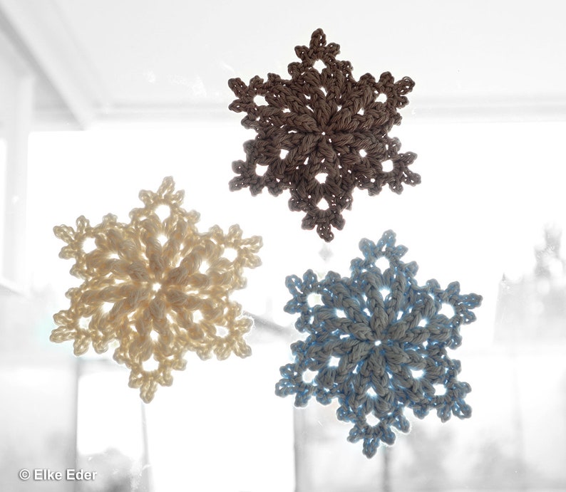 May include: Three crocheted snowflakes, one beige, one white, and one blue, hanging on a window.