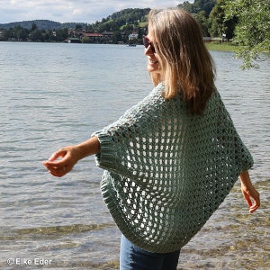 May include: A light blue crochet cardigan with a lacy openwork design. The cardigan is worn by a person standing near a lake.