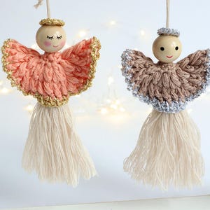 Könnte beinhalten: Zwei gehäkelte Engel-Anhänger mit Holzköpfen und weißen Garnquasten. Ein Engel ist pfirsichfarben mit goldenen Verzierungen und der andere ist braun mit silbernen Verzierungen.