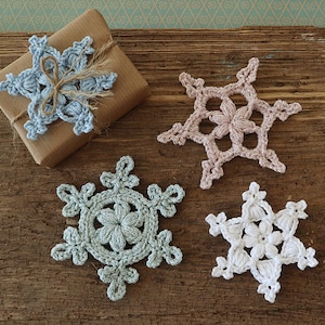 May include: Four crocheted snowflakes in different colours: light blue, pink, white, and light blue. The snowflakes are arranged on a rustic wooden surface.
