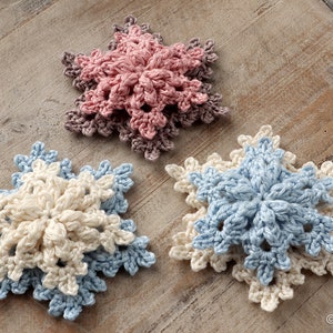 May include: Three crocheted snowflakes in different colors: pink, white, and blue. The snowflakes are layered on top of each other, creating a three-dimensional effect.