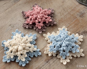 May include: Three crocheted snowflakes in different colors: pink, white, and blue. The snowflakes are layered on top of each other, creating a three-dimensional effect.