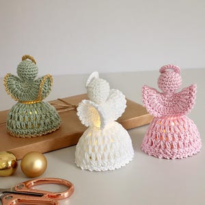May include: Three crocheted angel figurines in green, white, and pink. Each angel has a halo and a skirt-like body, with the green angel adorned with gold trim. The angels are lit from within, creating a soft glow.