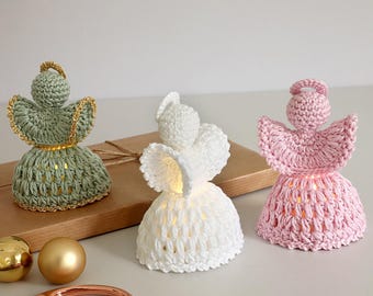 May include: Three crocheted angel figurines in green, white, and pink. Each angel has a halo and a skirt-like body, with the green angel adorned with gold trim. The angels are lit from within, creating a soft glow.
