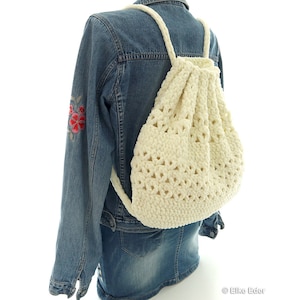 Könnte beinhalten: Ein weißer gehäkelter Rucksack mit einem weißen Kordelzug. Der Rucksack wird von einer Person getragen, die eine Jeansjacke trägt.