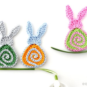 May include: Three crocheted Easter bunny ornaments with spiral designs. The bunnies are in pink, light blue, and gray with green, orange, and green spiral designs, respectively.