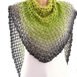 Puede incluir: Un chal hecho a mano, de tejido abierto, en un degradado de colores. El chal pasa de un verde lima brillante en el cuello a tonos de gris y negro en los bordes. La forma triangular y el diseño de tejido abierto crean un aspecto delicado y aireado.