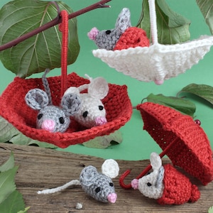 May include: Four crocheted mice, each holding a red and white crocheted umbrella. The mice are gray and white with pink noses. The umbrellas are hanging from a branch.