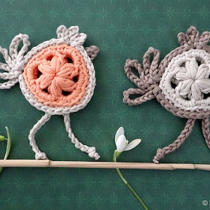 May include: Two crocheted birds with flower-shaped centers, one in peach and one in beige, perched on a branch with white snowdrop flowers.
