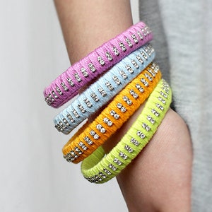 May include: Five colorful bracelets with rhinestones. The bracelets are made of yarn and are stacked on a person's wrist. The bracelets are pink, light blue, yellow, and green.