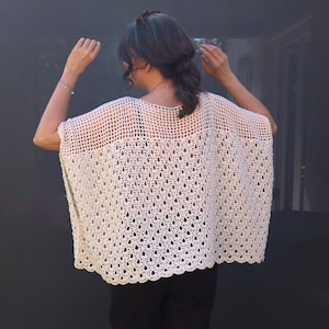 May include: A cream-colored, open-weave crochet top with a scalloped hem. The top has a loose, flowing design and a detailed floral pattern. The garment is worn over a black top and is suitable for warm weather.