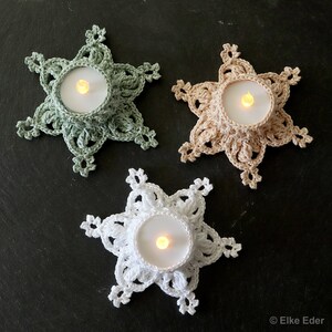 Puede incluir: Tres portavelas de copo de nieve de ganchillo en verde, beige y blanco con velas de té dentro.