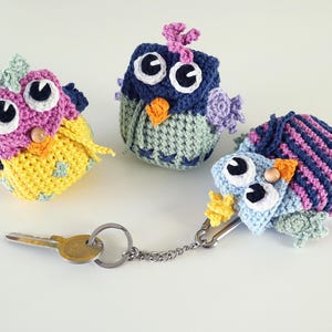 Op de afbeelding: Drie kleurrijke gehaakte uil sleutelhangers. Eén is geel, roze en blauw, een andere is blauw en groen, en de derde is blauw en roze gestreept. Elke uil heeft grote witte en zwarte ogen en een houten kralen neus. Een sleutel en ketting zijn bevestigd.