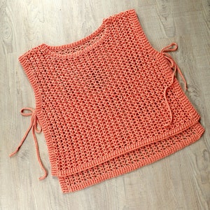 Könnte beinhalten: Ein korallenfarbenes Häkeltop mit lockerem, offenem Webmuster. Das Top hat zwei lange Bänder, die an den Seiten gebunden werden können.