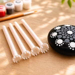 Op de afbeelding: Vier lichtroze dotting tools met een cirkelvormige kop liggen op een houten oppervlak. Rechts een zwart beschilderde steen met witte en grijze stippenpatronen. Kleine verfpotjes en een penseel op de achtergrond.