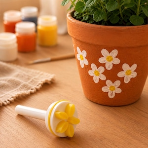May include: A white and yellow plastic flower-shaped craft stamp with a long handle, positioned near a terracotta clay pot painted with white and yellow daisy designs containing a green leafy plant, with small paint jars in the background.