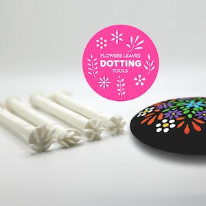 May include: Five white plastic dotting tools with different flower and leaf designs. The tools are arranged in a row on a white surface. A pink circle with the text "Flowers Leaves Dotting Tools" is in the background. A black rock with a colorful flower and leaf design is on the right side of the image.