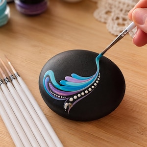 Puede incluir: Una piedra de río negra y lisa siendo pintada con un pincel de punta fina. El diseño presenta un patrón de remolinos en pintura azul claro y lavanda, acentuado por una serie de pequeños puntos blancos. Varios pinceles yacen cerca.