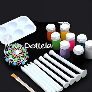 Puede incluir: Un kit de pintura de puntos con una roca negra y blanca con puntos de colores, una bandeja blanca con seis pozos, ocho pequeños frascos de pintura de varios colores, siete herramientas de punteado blancas y un pincel de madera. El kit está sobre una superficie negra con la palabra "Dottela" impresa en blanco.