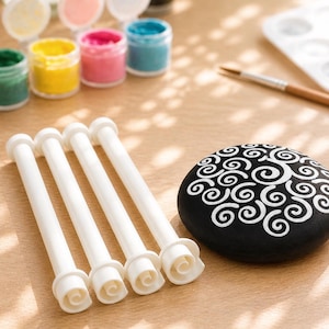 Peut inclure: Un ensemble de fournitures artistiques comprenant de petits pots de peinture de différentes couleurs, des outils blancs, un pinceau et une pierre noire peinte avec des motifs tourbillonnants blancs. Les articles sont disposés sur une surface en bois clair.
