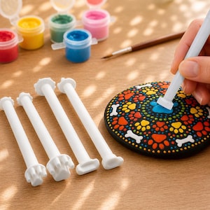 May include: A craft scene with colorful paint pots, a paintbrush, and white dotting tools. A hand is using a dotting tool to paint a blue dot on a black stone decorated with paw prints and bone shapes.