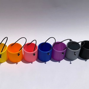 May include: A set of eight colorful, small, plastic buckets with black handles. The buckets are arranged in a row from left to right, starting with yellow, orange, pink, blue, purple, gray, black, and ending with white.