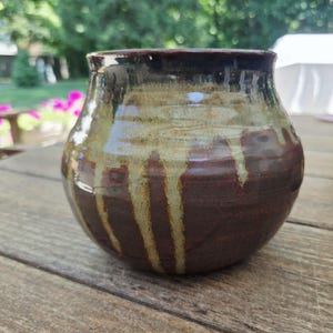 May include: A handcrafted ceramic vase with a rounded body and a wide opening. The vase features a dark brown base, transitioning to a tan and brown midsection, and a dark brown rim. The glaze has a dripped effect, adding visual interest.