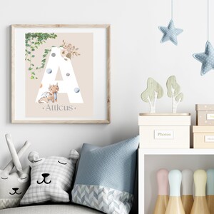 Könnte beinhalten: Gerahmter Druck mit einem großen weißen Buchstaben "A", einer Aquarell-Illustration eines Drachen und dem Namen "Atticus". Das Design umfasst florale und pflanzliche Elemente auf beigem Hintergrund. Der Rahmen ist aus hellem Holz.