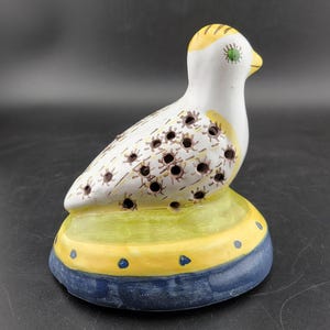 May include: A ceramic bird figurine with a white body, yellow accents, and brown spots. The bird has a green eye and is perched on a yellow and blue base.