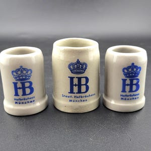 Könnte beinhalten: Drei cremefarbene Schnapsgläser aus Keramik. Jedes Glas zeigt ein blaues Kronen-Design über den blauen Buchstaben "HB" und den Worten "Hofbräuhaus München". Die Gläser sind zylindrisch mit leicht ausgestelltem Boden.