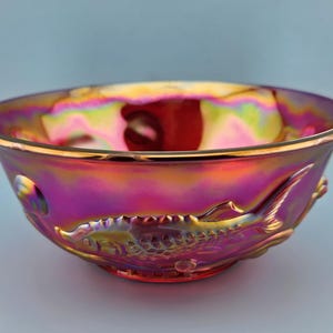 May include: Iridescent glass bowl with a fish design. The bowl has a vibrant color scheme of pink, gold, and purple. The fish is embossed on the side of the bowl. The rim of the bowl has a gold trim.
