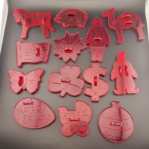 May include: A collection of thirteen red translucent plastic cookie cutters in various shapes including a horse, pig, camel, Santa Claus, American flag, poinsettia, butterfly, shamrock, dollar sign, clown, egg with Happy Easter text, carriage, and ornament.