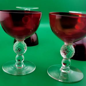 May include: Two stemmed glasses featuring deep red, bowl-shaped tops and clear glass bases. The stems are characterized by a central, spherical, textured knob with a diamond-patterned surface, positioned above a smaller smooth sphere and a flat base.