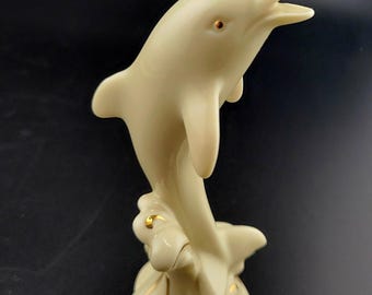 May include: A white porcelain figurine of a dolphin leaping out of the water. The dolphin is decorated with gold accents and is set on a base that resembles a wave.