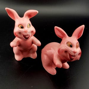 May include: Two pink ceramic bunny figurines with painted details. The bunnies have large eyes, a wide smile, and a pink tongue sticking out.