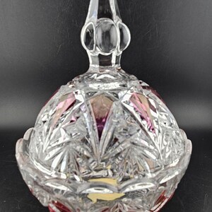 May include: A clear, cut-glass candy dish with a lid and three small feet. The dish has a round base with a red rim and a clear, pointed finial on the lid. The glass is intricately cut with a starburst pattern.