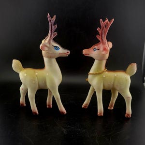 May include: Two vintage plastic reindeer figurines. The reindeer are cream-colored with pink antlers and accents. One has a gold-colored chain necklace. The reindeer have blue eyes and are standing on four legs. The background is black.