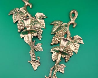 May include: Two brass wall sconces, each featuring a grape vine design with leaves and bunches of grapes. The sconces have a candle holder at the center. The background is a solid green color.