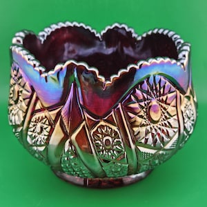 Puede incluir: Un cuenco decorativo de vidrio iridiscente con borde festoneado. El cuenco presenta un interior rojo oscuro y un exterior texturizado con patrones geométricos y florales. El vidrio refleja un arcoíris de colores, incluyendo púrpura, verde y dorado.