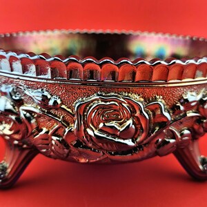 Puede incluir: Cuenco de vidrio iridiscente adornado con un diseño de rosas. El cuenco tiene un borde festoneado y se apoya en tres patas curvas. El patrón de rosas y hojas está en relieve en los laterales. El color del cuenco cambia de rojo a dorado.