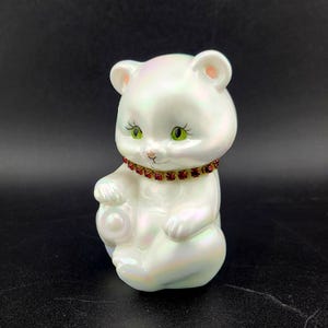 May include: A pearlescent white ceramic bear figurine with green eyes and a red jewelled collar. The bear is in a seated position, holding a small, round object. The figurine has a glossy finish against a dark background.