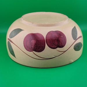 Puede incluir: Un cuenco de cerámica beige con un diseño pintado a mano de dos cerezas rojo oscuro y hojas verdes. El cuenco tiene forma redondeada y está sobre un fondo verde. Las cerezas tienen sombreado texturizado.