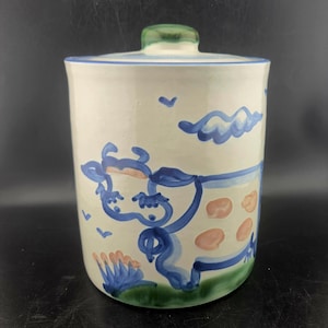 May include: A cylindrical ceramic canister with a lid featuring a hand-painted blue cow design on a cream background. The cow has peach-coloured spots, and the scene includes green grass, blue clouds, and small birds. The lid has a green knob.