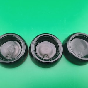 May include: Three black, glossy, circular objects with a recessed center and a small, stylized letter or symbol at the base. The objects are arranged in a row against a green background.